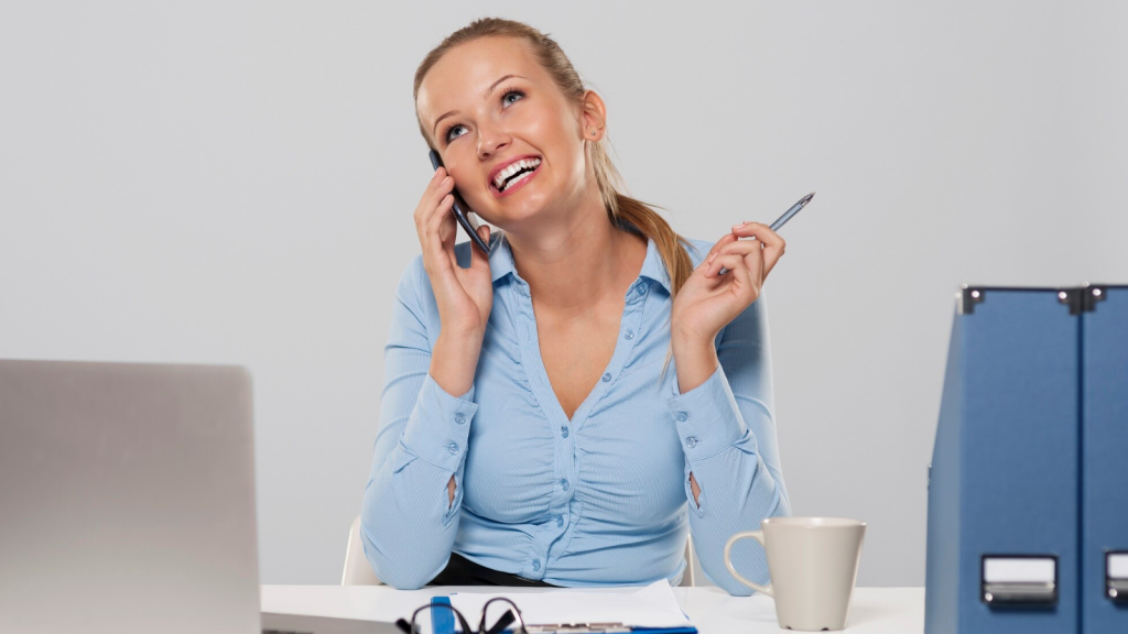 Smiling businesswoman in a blue blouse sitting at a desk, talking on a mobile phone while holding a pen. A laptop, documents, coffee mug, and office folders are visible on the desk. The image represents business phone features and team collaboration tools used in modern workplace communication.